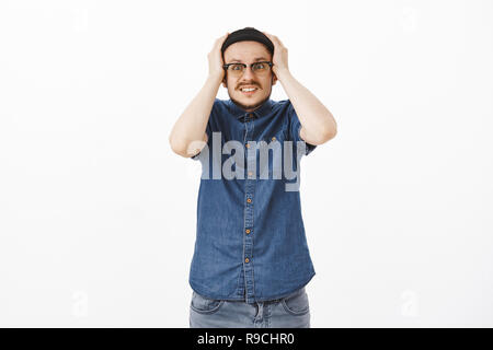 Choqué attrayant nerveux modèle masculin avec moustache et lunettes de beanie tenant les mains sur la tête de serrer les dents et regardant impatient en panique de l'appareil photo se souvenir qu'il a oublié de désactiver le fer Banque D'Images