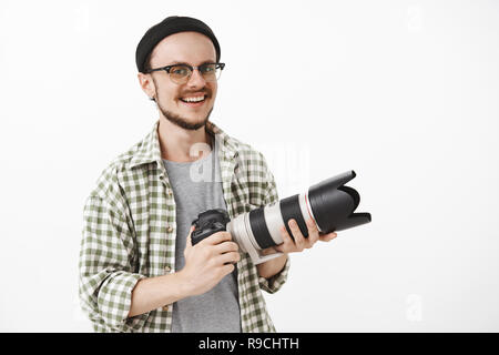 Jusqu'à la taille de balle de bonne énergie et d'enthousiasme à la mâle mature à lunettes et black beanie holding appareil photo professionnel et souriant avec joie de travailler en tant que journaliste ou photographe sur mur blanc Banque D'Images