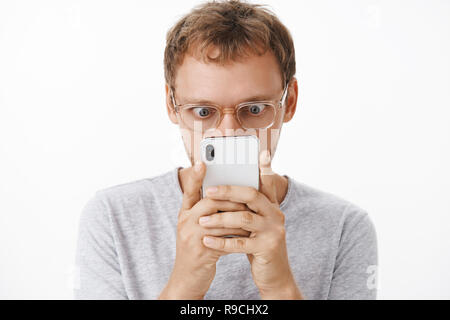 Close-up shot of amusé et diverti homme geek dans les verres holding smartphone près de visage yeux popping et fixant intrigué à l'écran de l'appareil lecture choquant message intéressant sur mur blanc Banque D'Images