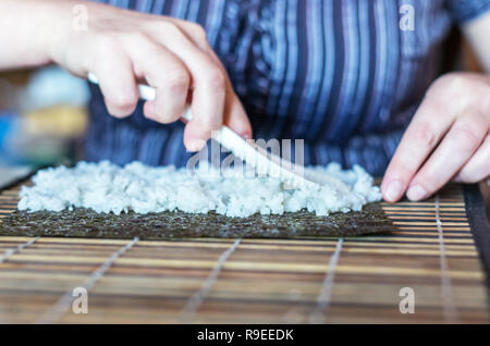 Faire de rouleaux de sushi à la maison : Répandre le riz blanc avec cuillère en plastique à travers la feuille d'algue Nori placé sur un tapis de bambou à la cuisine T Banque D'Images