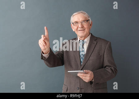 Senior male wearing eyeglasses studio isolés sur mur gris holding digital tablet à la recherche d'idées créatives dans internet à la caméra pointi Banque D'Images