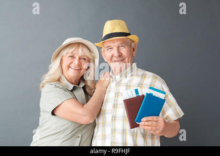 Couple de touristes studio isolés sur mur gris hugging tenant les passeports et les billets d'avion à la joyeuse souriante de l'appareil photo Banque D'Images