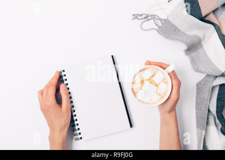 Woman's hands holding tasse de café avec des guimauves et ouvrir le bloc-notes en blanc sur fond blanc à côté de l'écharpe à damiers, vue du dessus. Mise à plat accueil chaleureux Banque D'Images