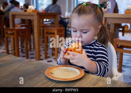 Beautiful happy young girl biting off grande tranche de pizza fraîche. Elle s'asseoir à chaise blanche dans un café et profiter de délicieux repas. Elle a les cheveux longs. Banque D'Images