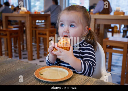 Beautiful happy young girl biting off grande tranche de pizza fraîche. Elle s'asseoir à chaise blanche dans un café et profiter de délicieux repas. Elle a les cheveux longs. Banque D'Images