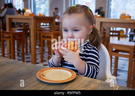 Beautiful happy young girl biting off grande tranche de pizza fraîche. Elle s'asseoir à chaise blanche dans un café et profiter de délicieux repas. Elle a les cheveux longs. Banque D'Images