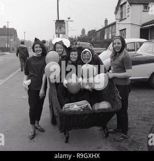 Entre 2 Lacs, 1967 Fête du village, un groupe de filles... habillés ils sont debout à l'extérieur dans une rue, trois d'entre eux assis dans un grand panier en osier sur un panier holding balloons, Buckinghamshire, Angleterre, Royaume-Uni. Banque D'Images