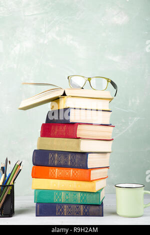 Une pile de vieux livres, des verres de couleur, de papeterie et d'une tasse de thé sur une table et un fond vert Banque D'Images