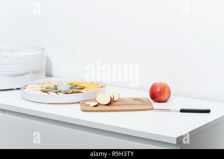 Sur une table blanche il y a une pomme rouge, une planche à découper, un couteau de cuisine, une bouteille et un bac avec des tranches de fruits. Banque D'Images