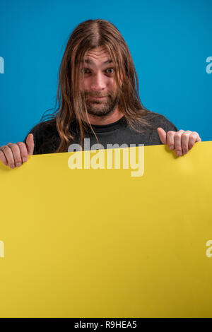 Portrait d'homme aux cheveux long faire une Drôle de tronche tout en tenant une bannière de publicité bleu. Copyspace disponible pour le message ou l'information Banque D'Images