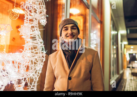 Young smiling man walking in city par Noël décoré en vedette la nuit et à la recherche à l'appareil photo. Nouveau concept de l'année Banque D'Images
