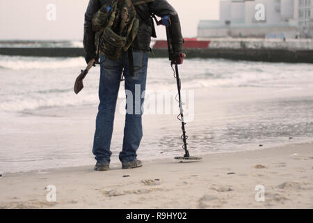 Digger noir. Homme avec détecteur de métal sur la plage à la recherche d'objets perdus sous le sable de l'océan, sur la mer. Banque D'Images