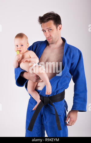 Karaté sportif homme en kimono bleu avec ceinture noir debout, tenant un bébé garçon et looking at camera Banque D'Images