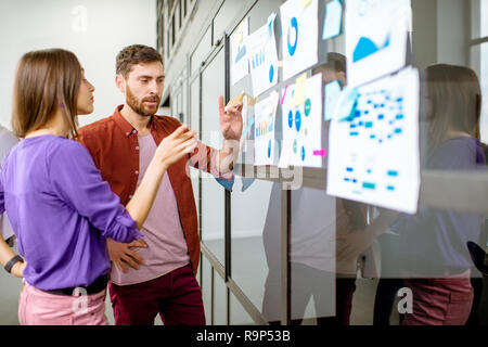 Jeune homme et femme vêtue de désinvolture travailler avec quelques statistiques sur le mur de verre dans le bureau Banque D'Images