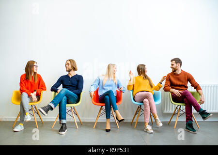 Portrait d'un jeune gens habillés nonchalamment assis dans une rangée sur le chaises colorées sur le mur blanc Banque D'Images