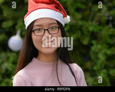 Jolie Thai teen girl avec un Santa Claus hat déclinée pour l'appareil photo en face d'un arbre de Noël décoré. Banque D'Images