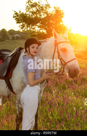 Prendre soin des animaux, l'amour et l'amitié concept. Jeune Jockey girl petting et serrant cheval blanc dans le coucher du soleil du soir. Serveur Sun flare Banque D'Images