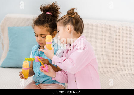 Adorable enfants multiethnique assis sur un canapé et en soufflant des bulles de savon Banque D'Images