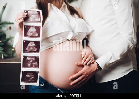 Heureux mari et femme posant ensemble. Jolie pregnant woman holding ultrasound photos du futur bébé. L'homme en chemise blanche tenant la main sur le ventre. De Banque D'Images