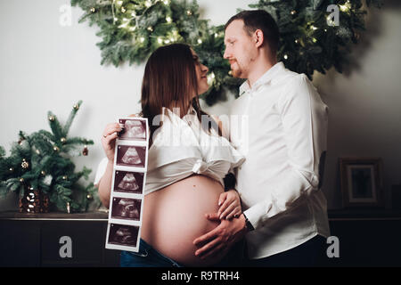 Heureux mari et femme posant ensemble. Jolie pregnant woman holding ultrasound photos du futur bébé. L'homme en chemise blanche tenant la main sur le ventre. De Banque D'Images