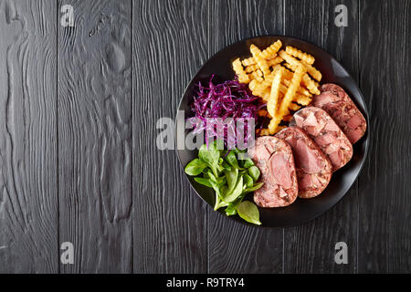 Delicious tranches de terrine de langue de boeuf et la viande servie avec frites, feuilles vertes et rouges salade de choux sur une plaque noire sur une table en bois, vue Banque D'Images