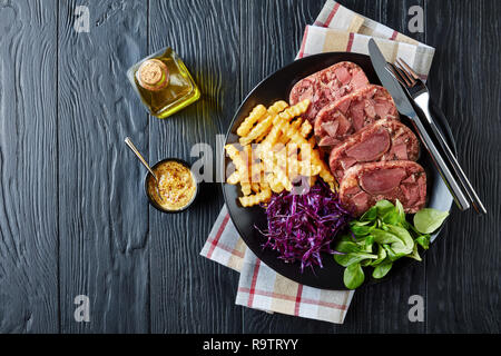 Vue de dessus de la langue de boeuf en tranches de viande et de l'aspic servis avec frites, feuilles vertes et rouges salade de choux sur une plaque noire sur une table en bois wit Banque D'Images