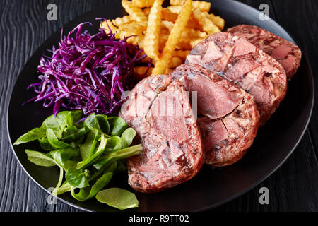 La langue de boeuf en tranches de viande et de l'aspic servis avec frites, feuilles vertes et rouges salade de choux sur une plaque noire sur une table en bois, vue de dessus, cl Banque D'Images