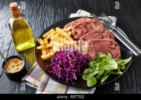 La langue de boeuf en tranches l'aspic servis avec frites, feuilles vertes et rouges salade de choux sur une plaque noire sur une table en bois avec de la moutarde dans un bol, vue Banque D'Images