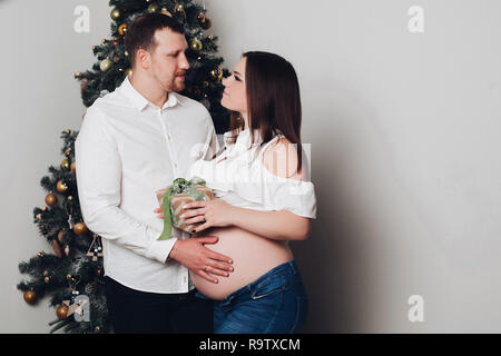 Cheerful mari et femme debout, posant près de l'arbre de Noël et de coffrets cadeaux. L'homme en chemise blanche tenant la main sur le ventre de la femme. Future maman avec na Banque D'Images