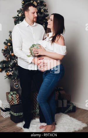 Cheerful mari et femme debout, posant près de l'arbre de Noël et de coffrets cadeaux. L'homme en chemise blanche tenant la main sur le ventre de la femme. Future maman avec na Banque D'Images