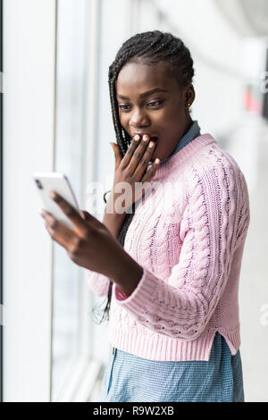 Jeune femme afro surpris choqué lire message de téléphone devant des fenêtres panoramiques Banque D'Images