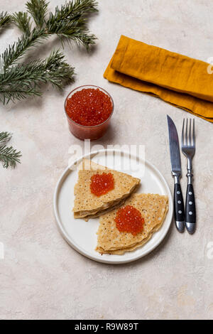 Crêpes minces avec du caviar rouge à plaque blanche avec pot de caviar rouge, fourchette, couteau, serviette et près de sapin à fond clair Banque D'Images