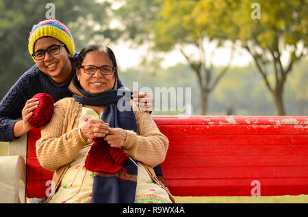 Heureux à la jeune femme indienne avec sa mère knitting sweater qui est assis sur un banc rouge dans un parc à New Delhi, Inde Banque D'Images