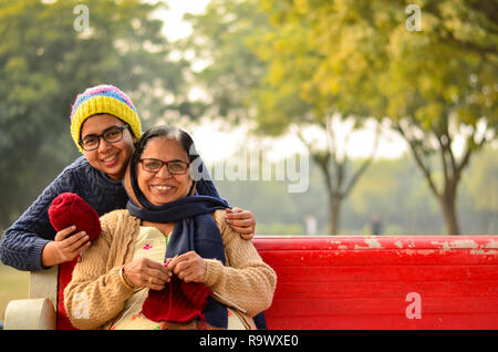 Heureux à la jeune femme indienne avec sa mère knitting sweater qui est assis sur un banc rouge dans un parc à New Delhi, Inde Banque D'Images