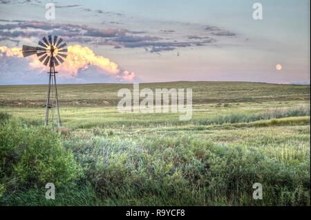 Un moulin à vent sur le Colorado's prairie à graminées courtes avec un fond de nuages éclairés par le soleil couchant et la pleine lune se lever. Banque D'Images