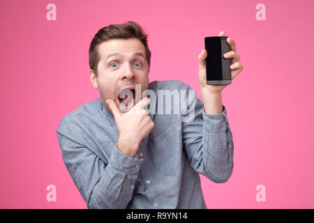 Handsome young man holding smartphone, montrant à l'appareil photo gadget, isolé sur fond rose. Banque D'Images
