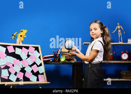 Jeune fille se tient par tableau noir avec des notes de couleur. Retour à l'école et l'enfance concept. Kid et fournitures scolaires sur fond de mur bleu. Écolière avec sourire globe tient dans ses mains Banque D'Images