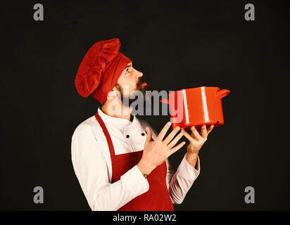 Concept de cuisine professionnelle. Cocotte rouge Chef détient ou casserole. Cook avec sérieux en bourgogne hat et de l'aire de recherche et est titulaire d'casserole. Homme à barbe est titulaire d'ustensiles sur fond noir. Banque D'Images