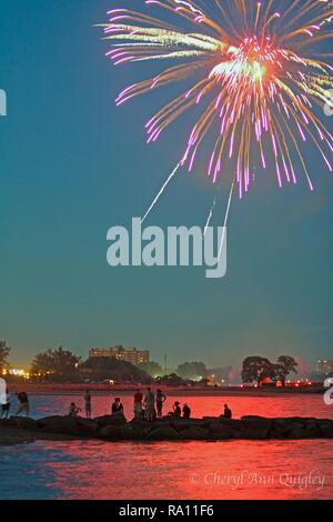 Groupe de personnes artifice à partir d'une jetée. La réflexion d'artifice s'allume en rouge dans l'eau autour d'eux. Banque D'Images