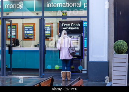 Une personne dans un manteau rose avec de longs cheveux blonds portant des bottes d'hiver en daim fourrure à l'aide d'un guichet automatique situé dans la fenêtre d'une boutique à Bath eurochange Banque D'Images