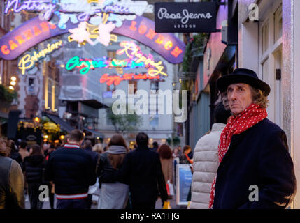 Homme avec écharpe rouge & hat s'appuie contre le mur à la recherche en tant que peuple marche par shopping sur Carnaby Street, en vertu de l'Installation de l'éclairage au néon Noël.2018 Banque D'Images