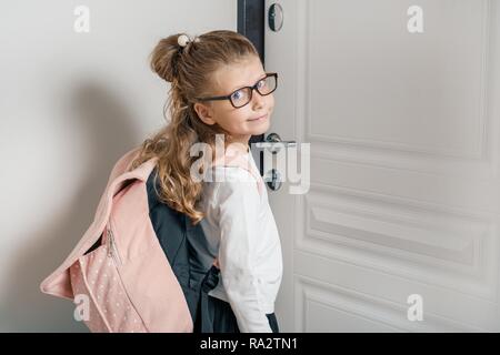Peu jolie fille 6, 7 ans avec sac à dos pour l'école. Jeune fille souriante, debout près de la porte de la chambre, enfant va à l'école. Banque D'Images