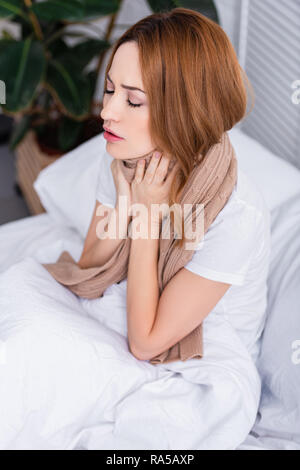 Portrait de femme malade de toucher le cou douloureux avec foulard et assis sur le lit à la maison Banque D'Images