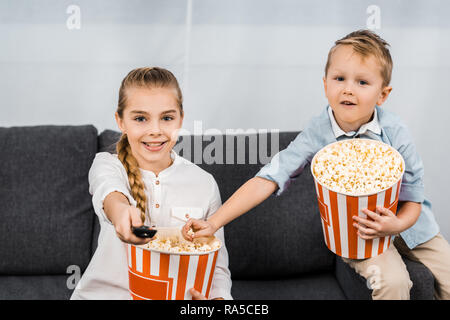 Smiling siblings sitting on sofa holding, seaux de pop-corn à rayures et des canaux en évolution par télécommande Banque D'Images