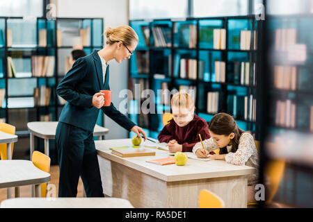 Jeune enseignante holding cup et à la recherche à étudier et à écrire en écolières library Banque D'Images