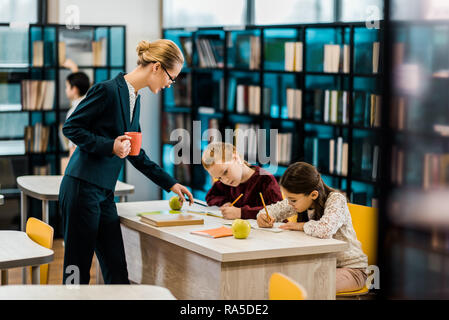 Jeune enseignante holding cup et à des écoliers à studying in library Banque D'Images