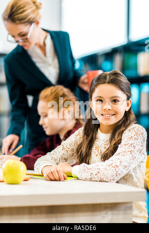Cute schoolkid dans eyeglasses smiling at camera while étudie avec camarade et enseignant dans la bibliothèque Banque D'Images