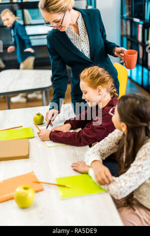 Souriante jeune enseignant dans les lunettes à des écoliers à studying in library Banque D'Images