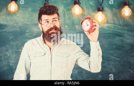 Enseignant dans les lunettes est titulaire d'un réveil. Hipster barbu est titulaire d'réveil, tableau sur toile de fond, l'espace de copie. Homme avec barbe et moustache sur le visage ennuyé à l'écoute d'alarme. Cloche de concept. Banque D'Images