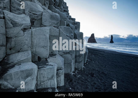 Falaise de colonnes de basalte le long de la plage de sable noir de Reynisfjara qui jouit, avec la mer de Reynisdrangar piles dans la distance, de l'Islande Banque D'Images